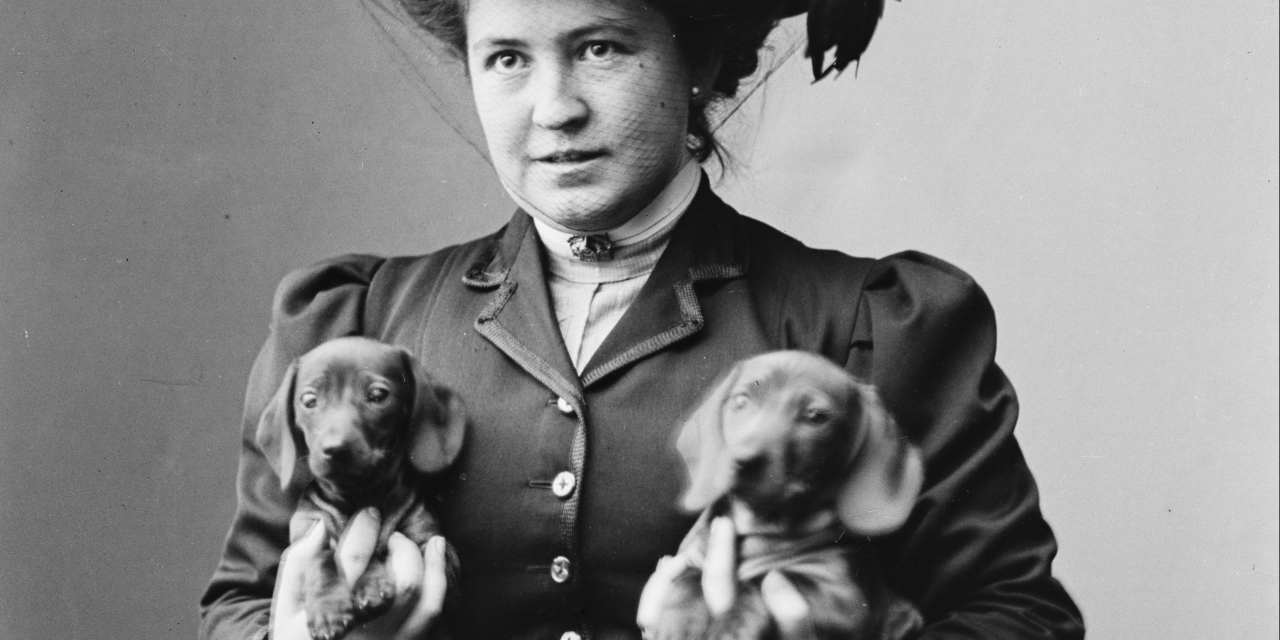 Fotografi af en kvinde i fin kjole og en stor hat med en gravhund under hver arm.