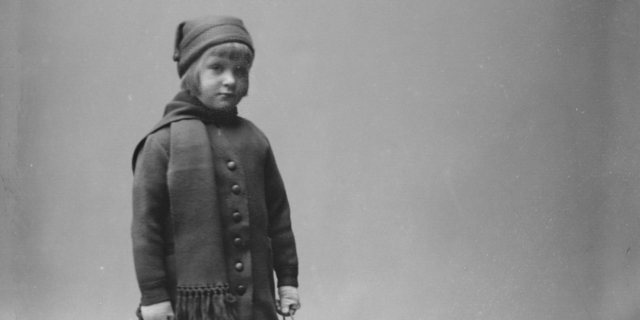 Portrætfotografi af en lille pige med hue og halstørklæde på og en kælk i hånden.