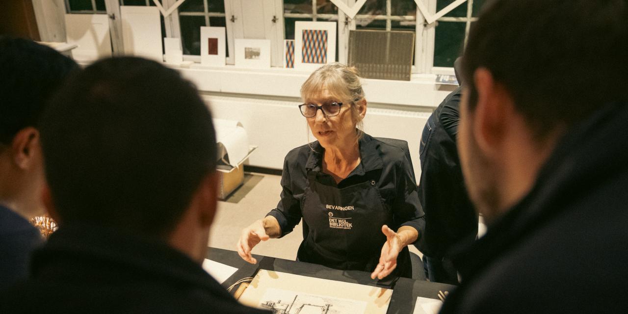 En medarbejder fra Det Kgl. Bibliotek sidder ved et bord og taler med gæster, der står rundt omkring hende.