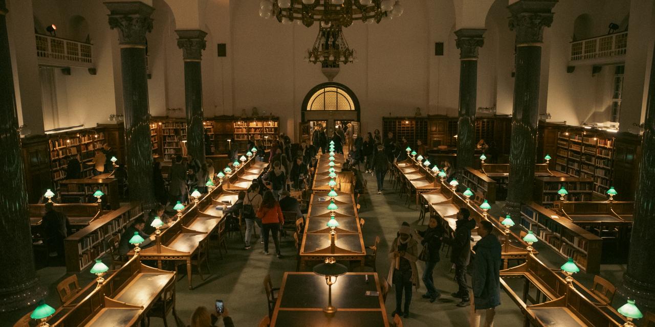 Gammel Læsesal oplyst i mild belysning med gæster, der går rundt i det store rum