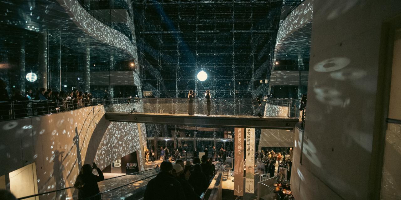 To sangere står i midten af Atriet og synger i skæret fra den store spejlkugle.