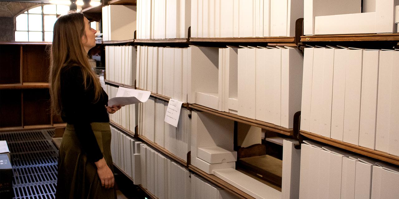 Kvinde står ved reoler fyldt med mapper med et stykke papir i hånden. Måske leder hun efter noget specifikt?