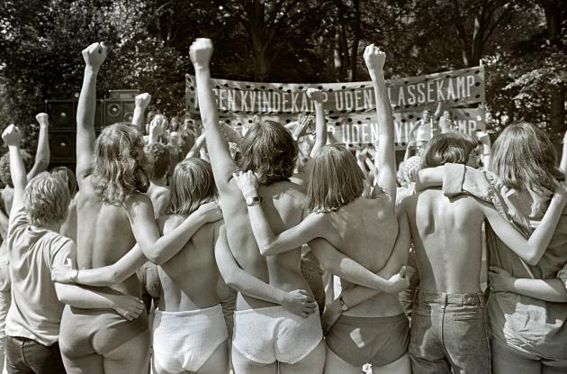 A group of half-naked people stand with their backs to the camera with arms either around each other or in the air