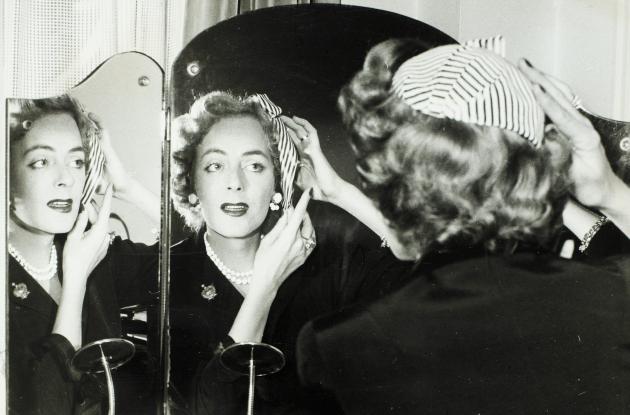 Christine Jorgensen straightens her hat in front of a dressing table with a mirror