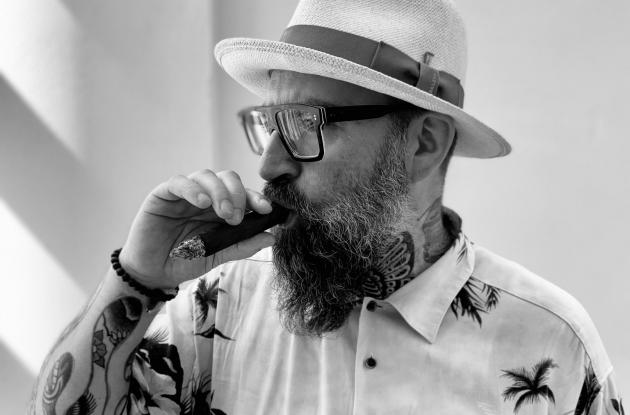 Sort-hvid billede af digteren Claus Høxbroe i sommerskjorte og hat, ryger en cigar.