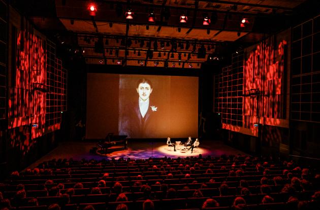 The Queen's Hall is bathed in red light over the audience, on stage three people are immersed in conversation and on the back wall is a large portrait of Marcel Proust