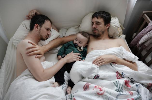 Two fathers lie in a bed with no shirts on with their small child in between them
