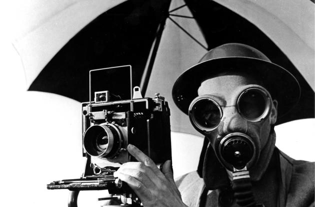 Black and white image of David E. Scherman with gas mask and camera on tripod.
