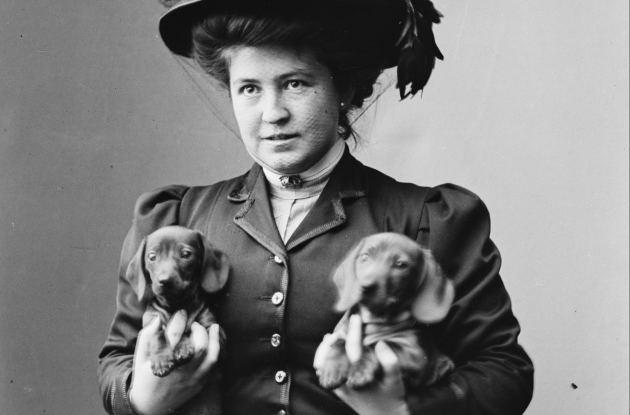 Fotografi af en kvinde i fin kjole og en stor hat med en gravhund under hver arm.