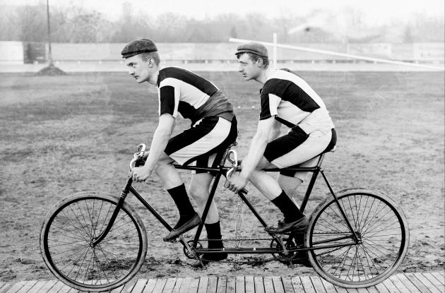 Photograph of two men in striped suits riding a tandem bicycle.