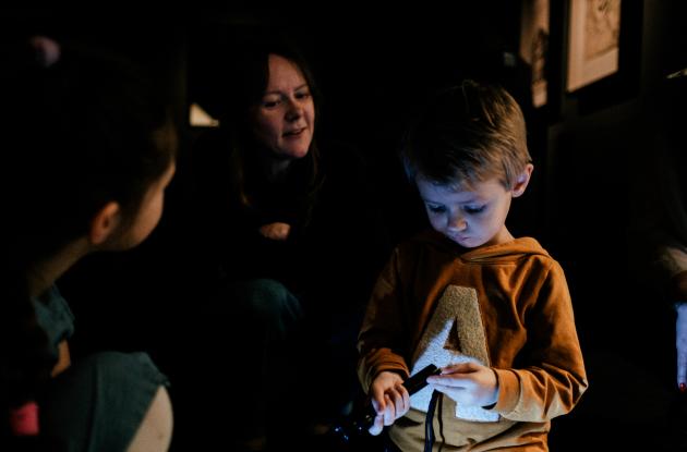 Children stand with an adult in a dark room, where the little boy stands with a flashlight in his hand.