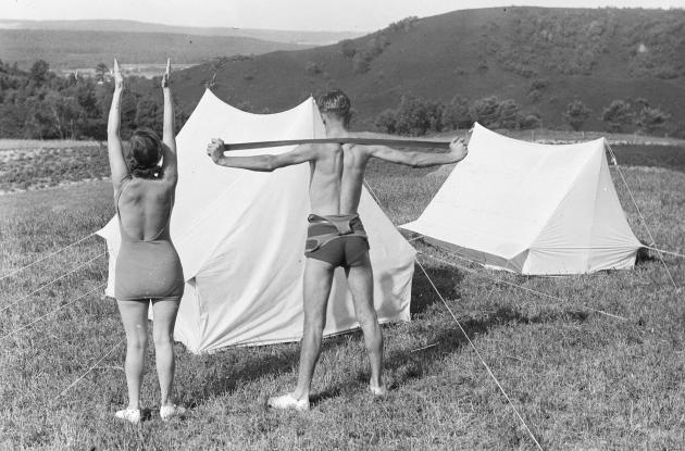 En mand og en kvinde der dyrker morgengymnastik i naturen ved nogle telte, fotograferet bagfra