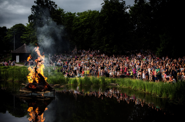 Sankt Hans bål på sø med en masse mennesker samlet på bredden.