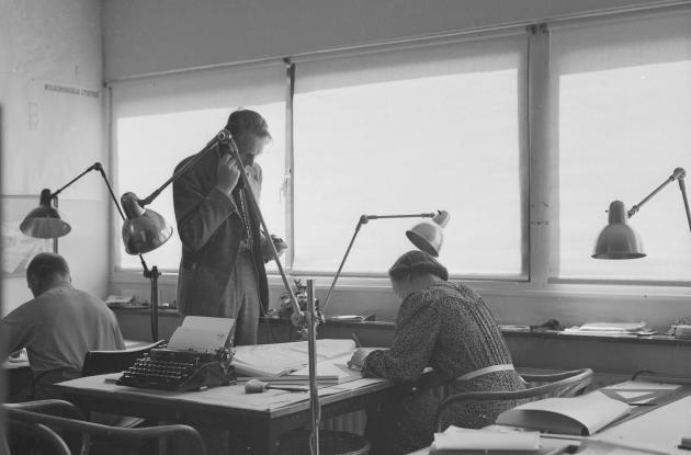 Mand taler i telefon stående på et kontor. Kvinde sidder ved skrivebord og skriver eller læser.