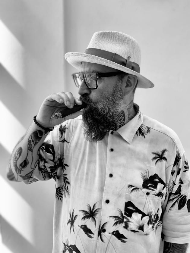 Sort-hvid billede af digteren Claus Høxbroe i sommerskjorte og hat, ryger en cigar.  