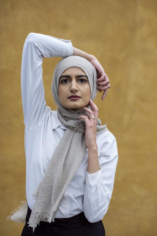 The poet Naiha Khiljee poses in white in front of an ocher yellow wall.