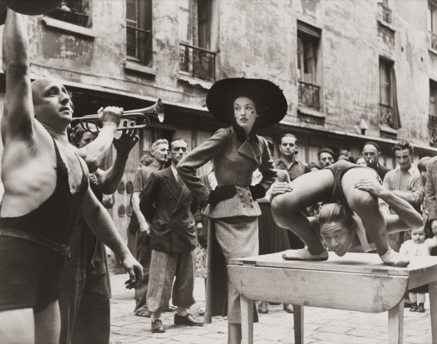 En model poserer på gaden, omringet af gadeperformere. En spiller trompet, en anden poserer i en hypermobil stilling på et bord. Modellen bærer et sæt med blazer og nederdel og en stor hat.