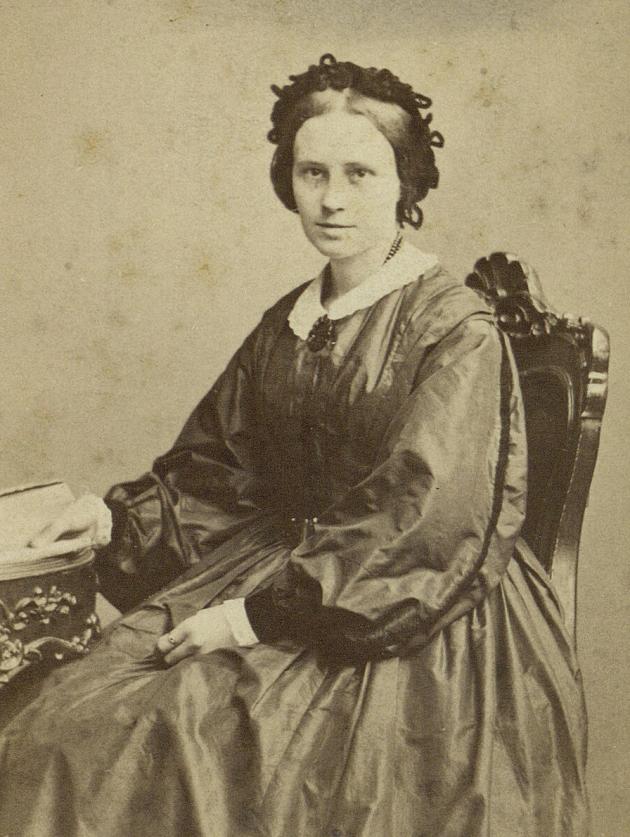 Mathilde Fibiger is sitting with a book in a nice dress. She has the other hand in her lap.