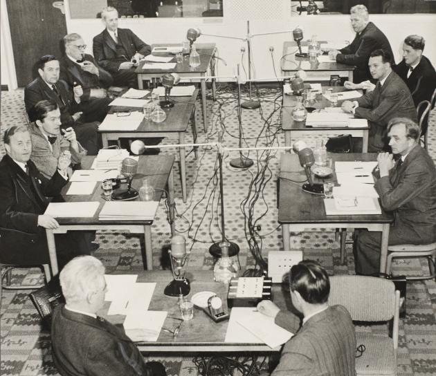 Several men sit at desks and speak into microphones