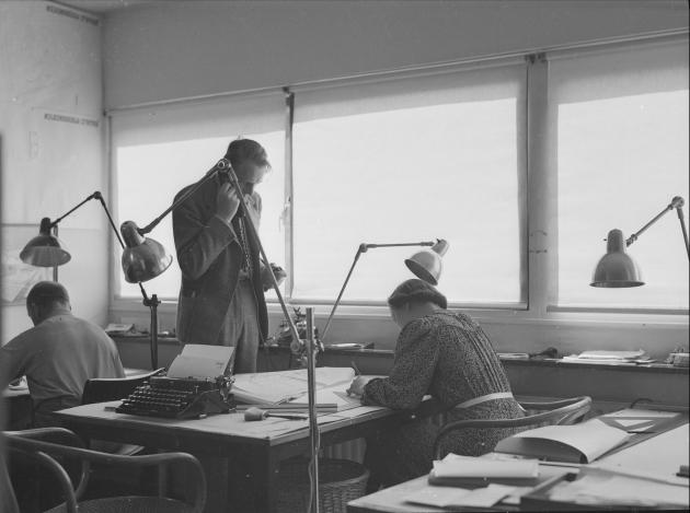 tre mennesker på et kontor. To sidder, en står op og taler i telefon.