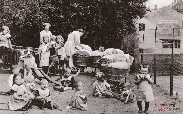 Vuggestue; to kvinder og en masse børn. Nogle af børnene er i barnevogne mens andre sidder på græsset.