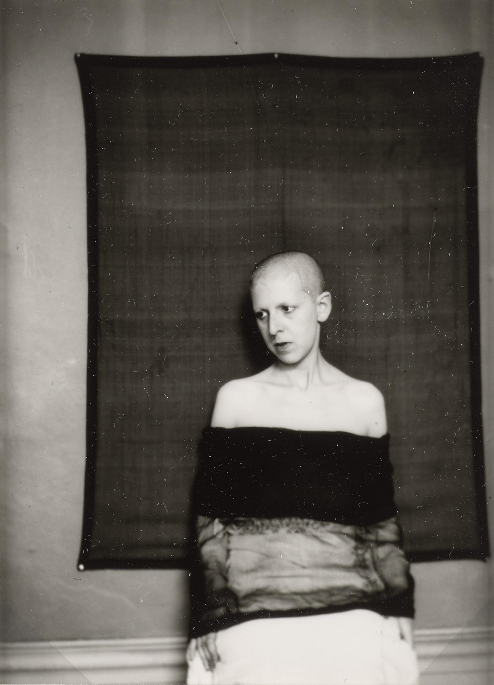 Selvportræt af Claude Cahun, der poserer foran et mørkt vægtæppe.