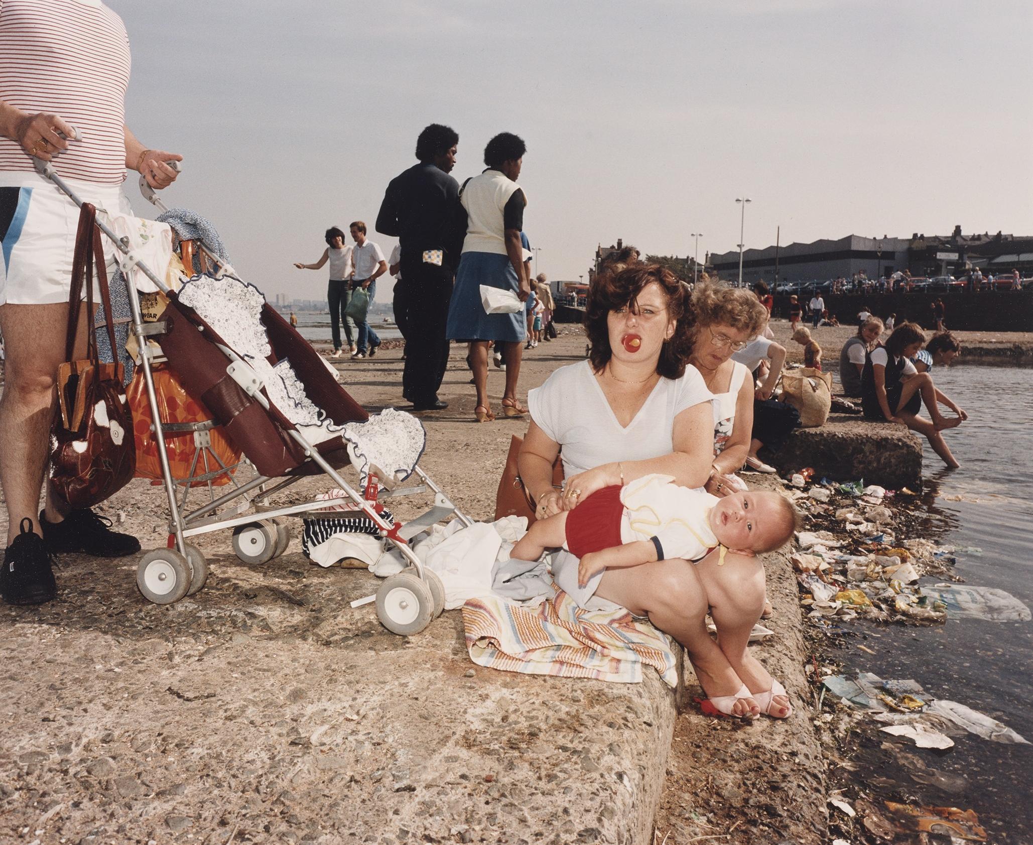 En kvinde sidder med en klapvogn og en baby ved noget vand, 1980erne