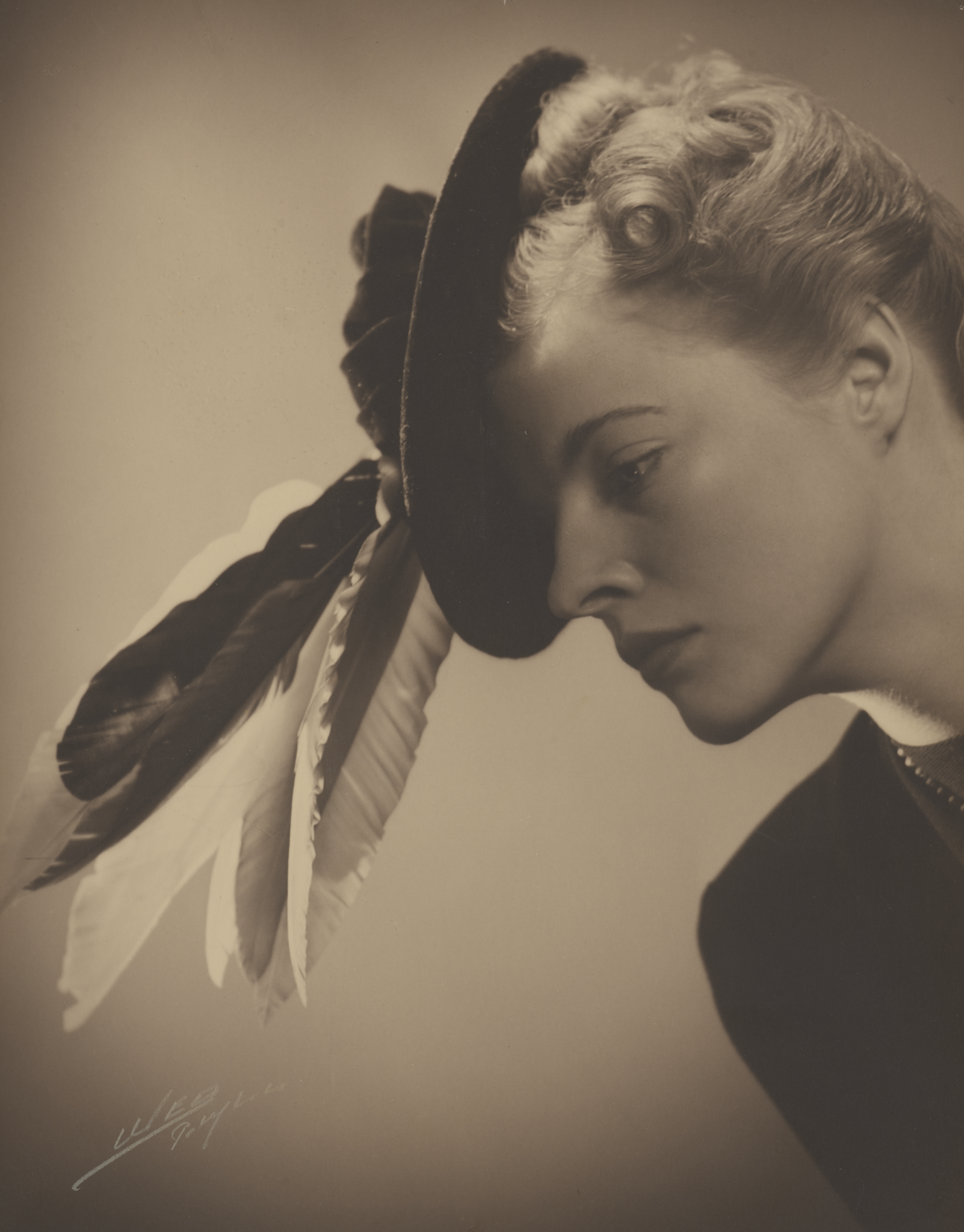 Kvinde kigger nedad og læner sig frem. Hendes hår er krøllet op, og hun bærer en fin hat med fjer i.