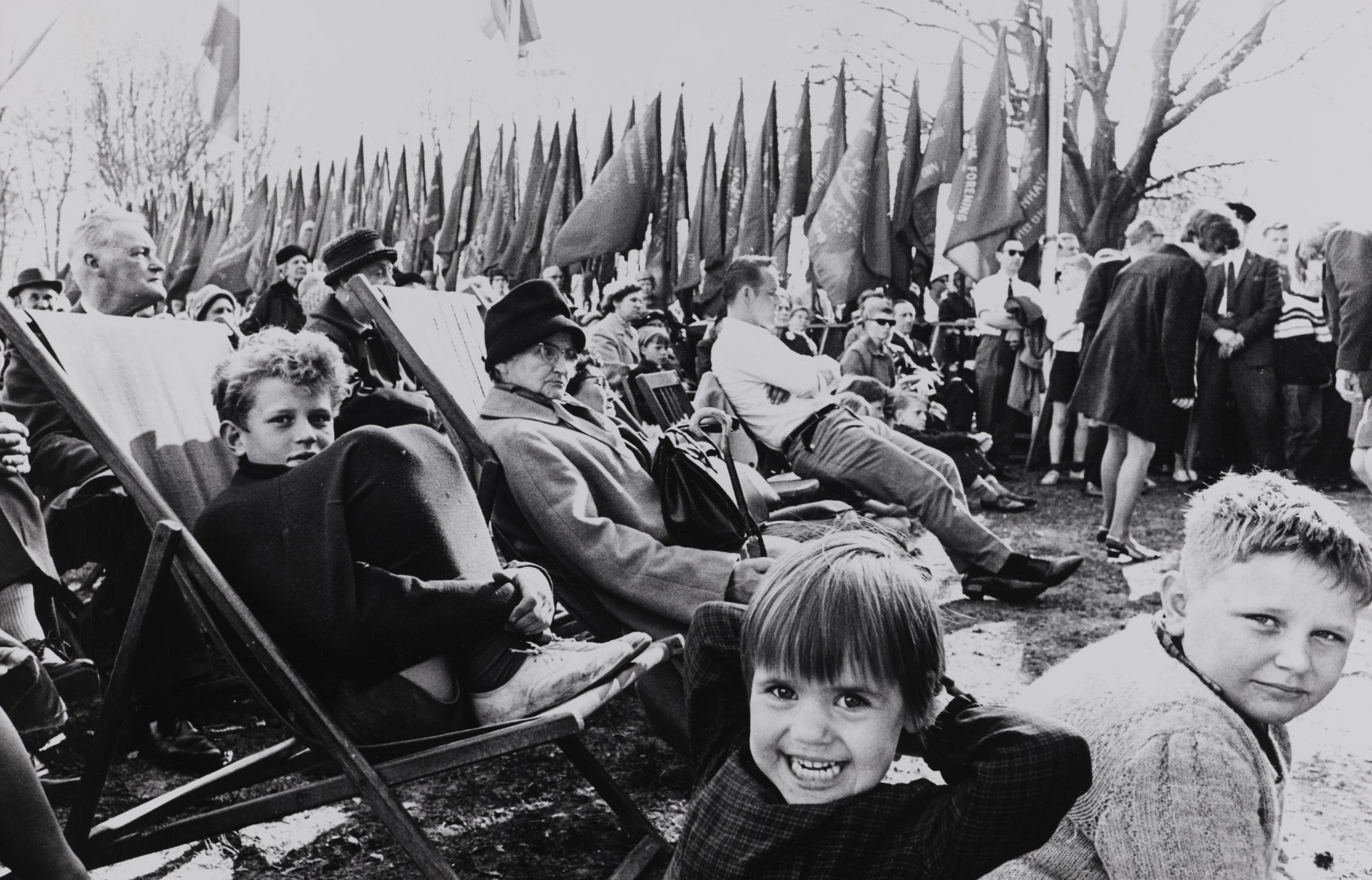 En gruppe mennesker sidder i stole foran en række af faner. Tre drenge i forgrunden smiler til kameraet.