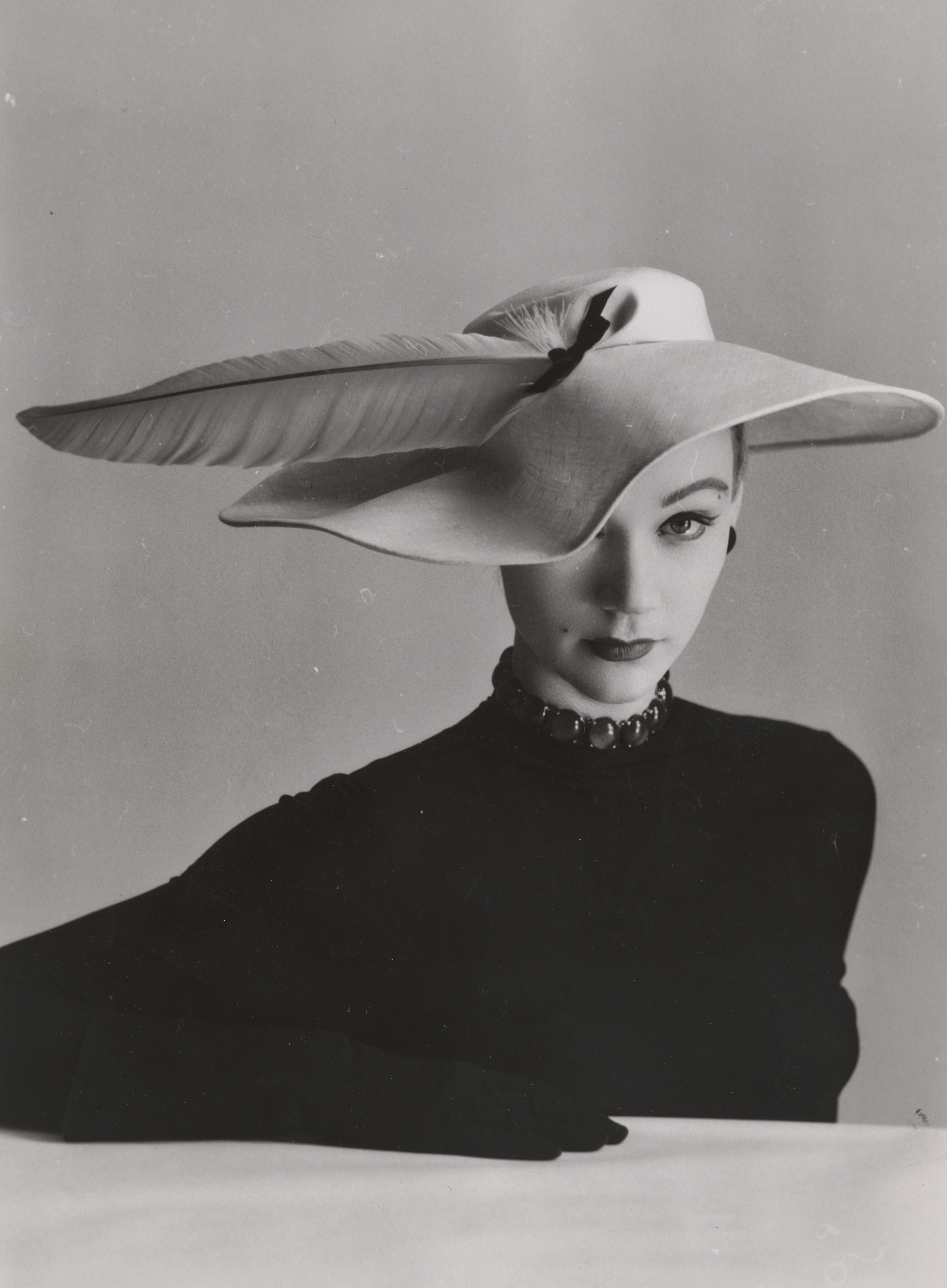 Kvinde med sort trøje og handsker sidder ved et bord. En hat med en enkel fjer skygger for hendes ene øje.