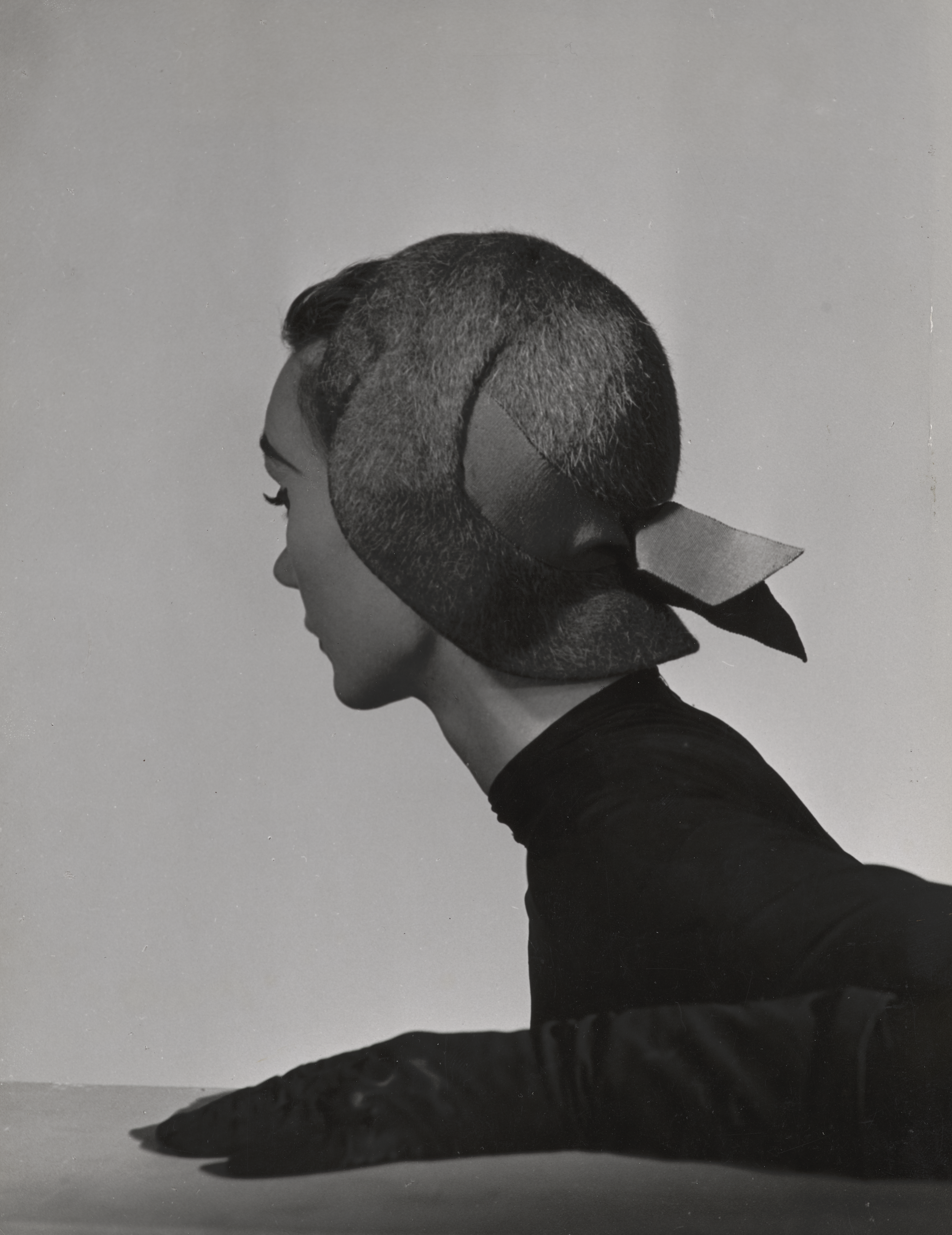 Kvinde med sort trøje og handsker sidder ved et bord. Hun ser væk fra kameraet, så man kan se hendes fine hat med sløjfe.