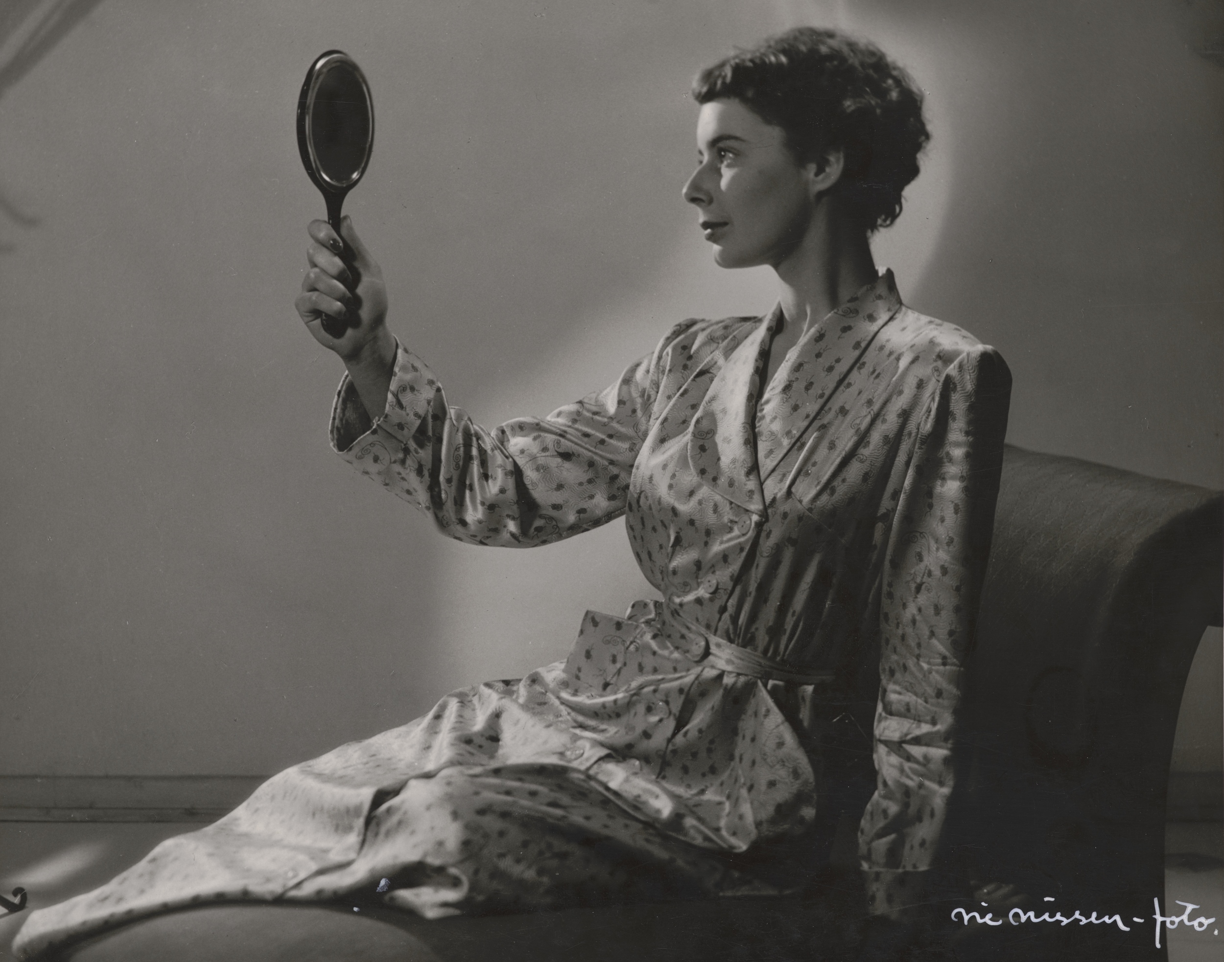 Kvinde ned og kigger sig selv i et lille håndholdt spejl. Hendes hår er sat op og bag hende oplyses en væg.