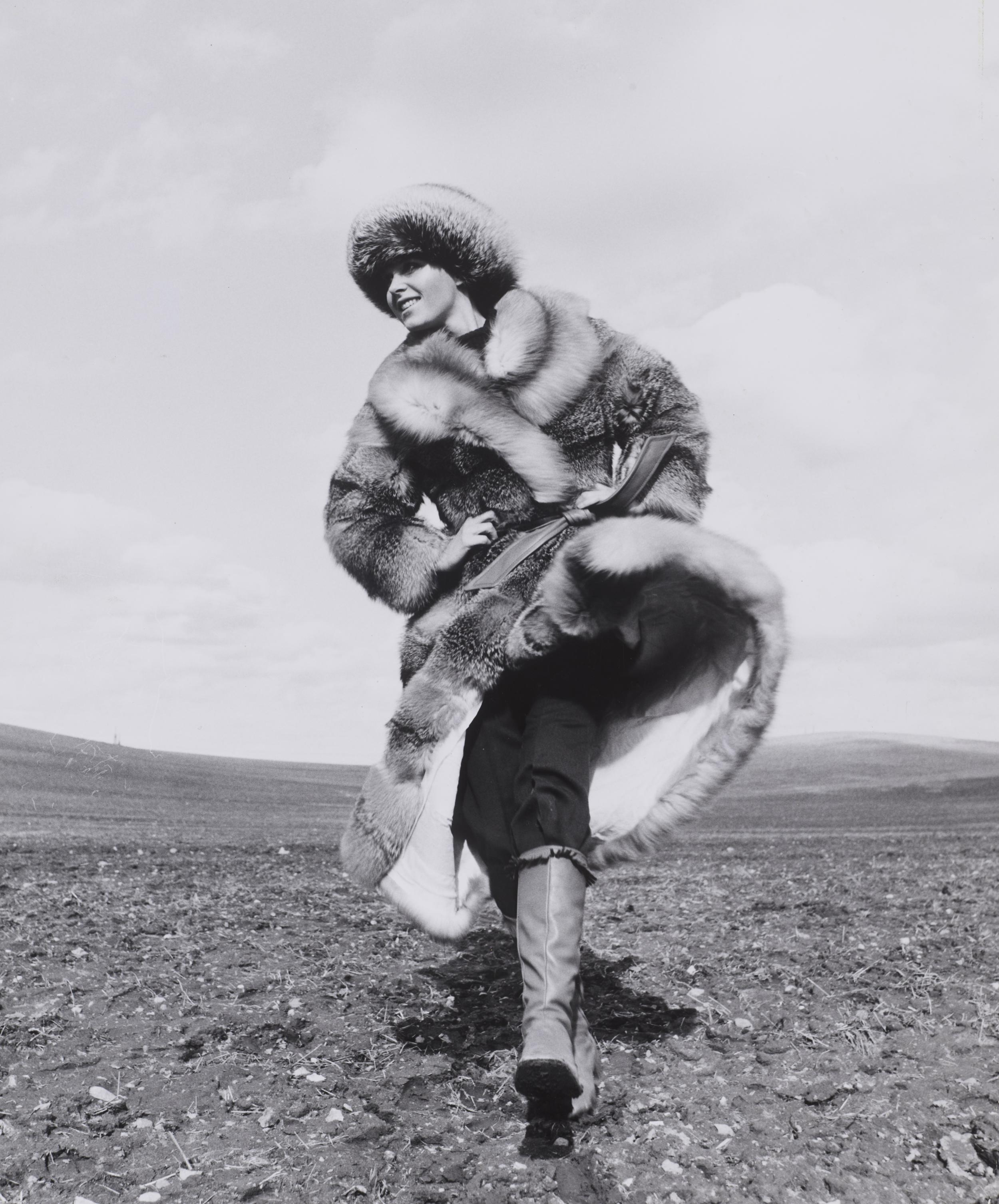 Lotte Freddie i rask gang med et øde landskab i baggrunden. Hun er iklædt en stor pelsfrakke, pelshue og skindstøvler.