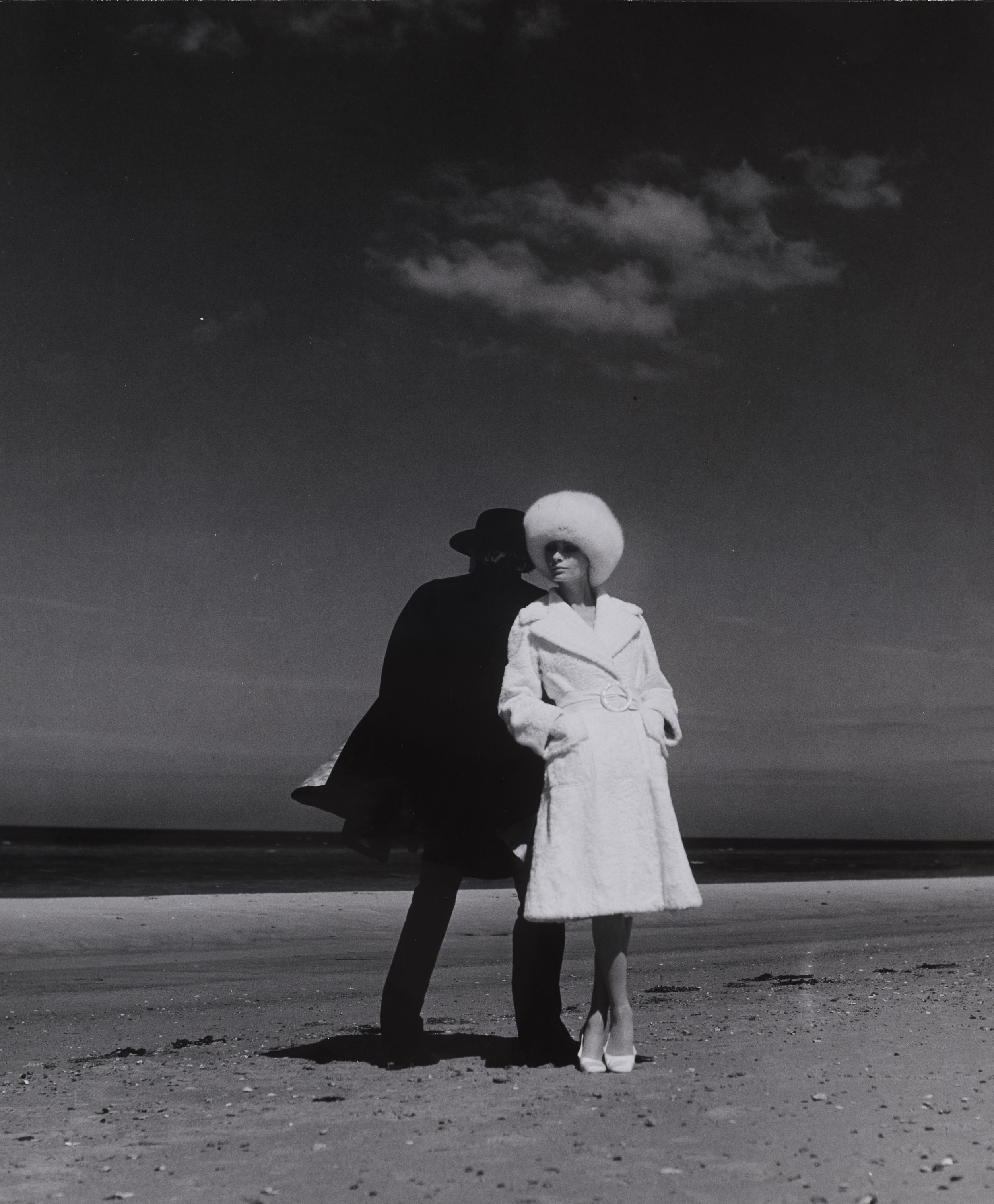 Kvinde poserer i frakke og pelshat på en strand. Bag hende er en mand henlagt i skygge.