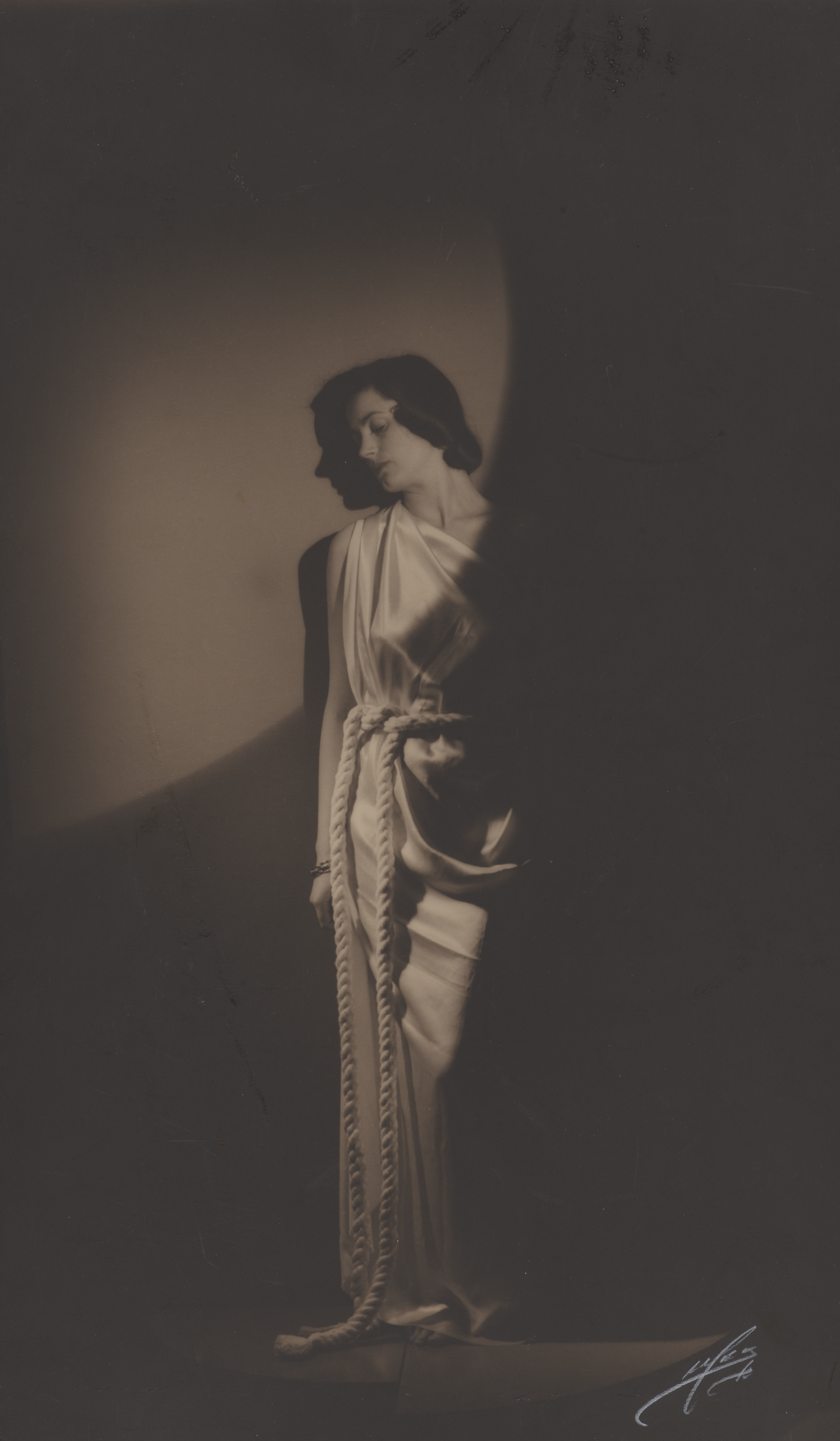 Kvinde står op ad en væg. Hun er klædt i en græsk-inspireret kjole med reb som bælte. Et enkelt lys skinner på hendes ansigt.