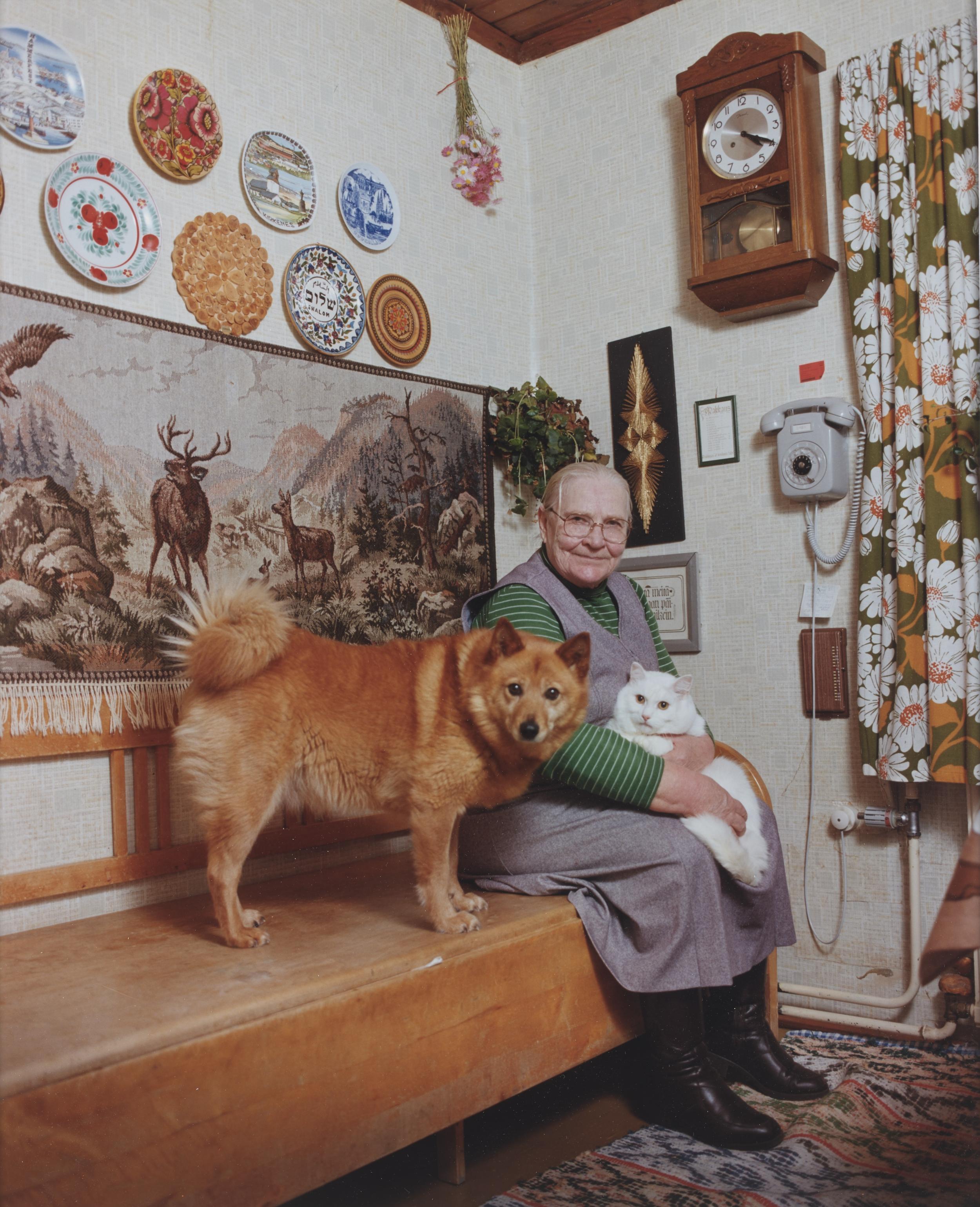 En ældre kvinde sidder i et dekoreret rum med en hvid kat på skødet og en brun hund ved siden af. Alle tre ser ind i kameraet