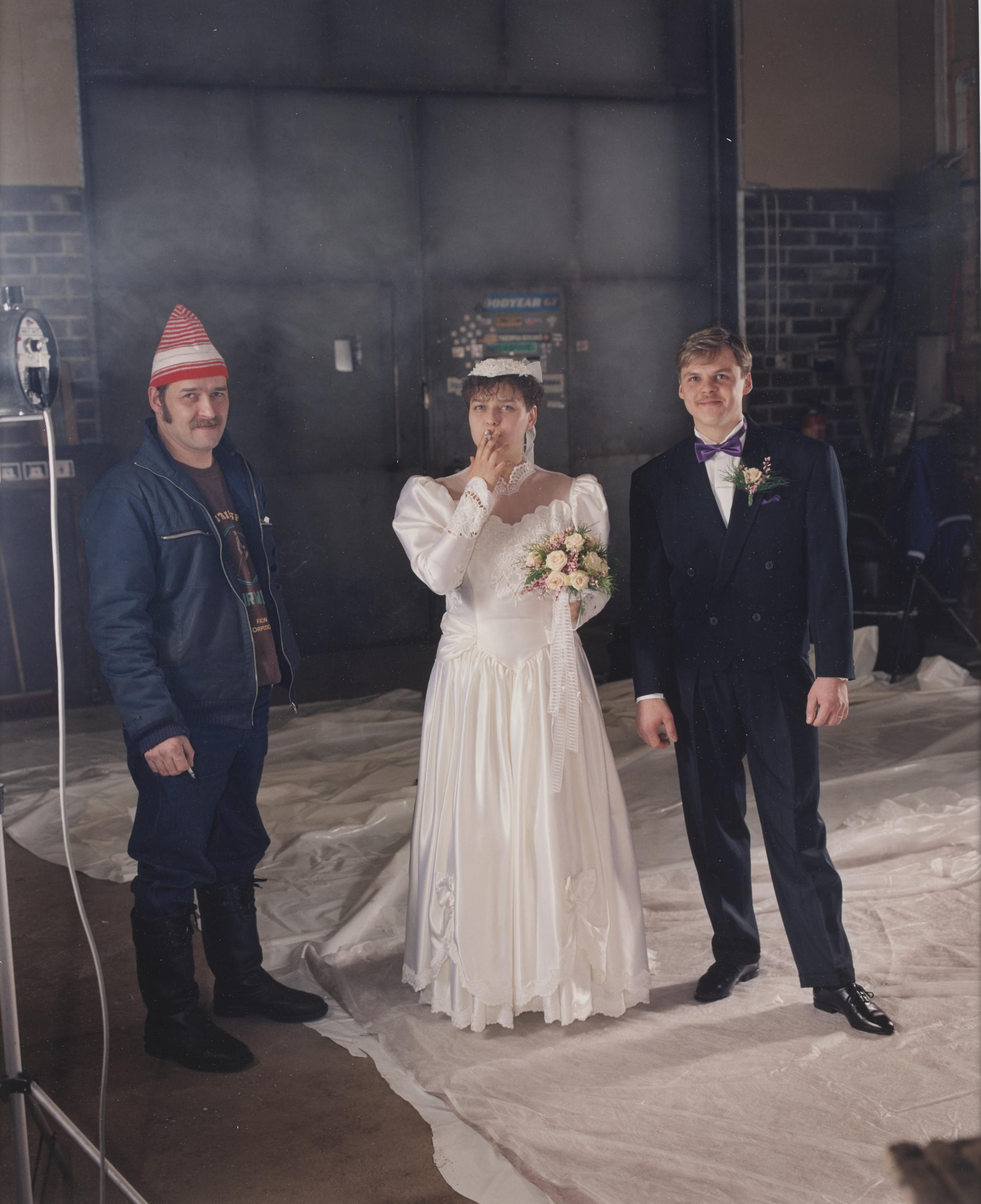Et brudepar står ved siden af en mand i denimjakke og rød hat
