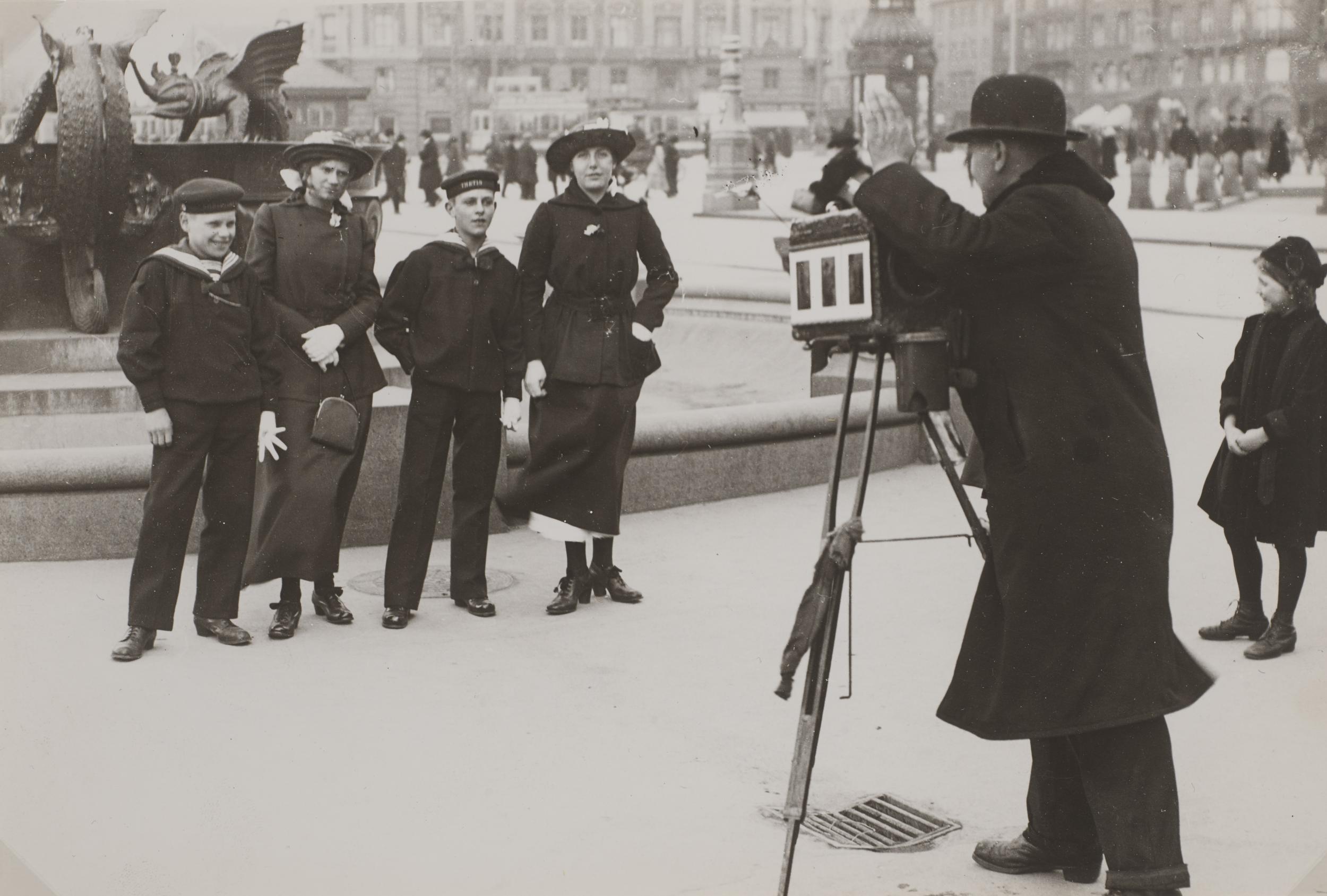 Fotograf med gammeldags kamera tager billede af fire mennesker foran et springvand. To kvinder og to mænd.