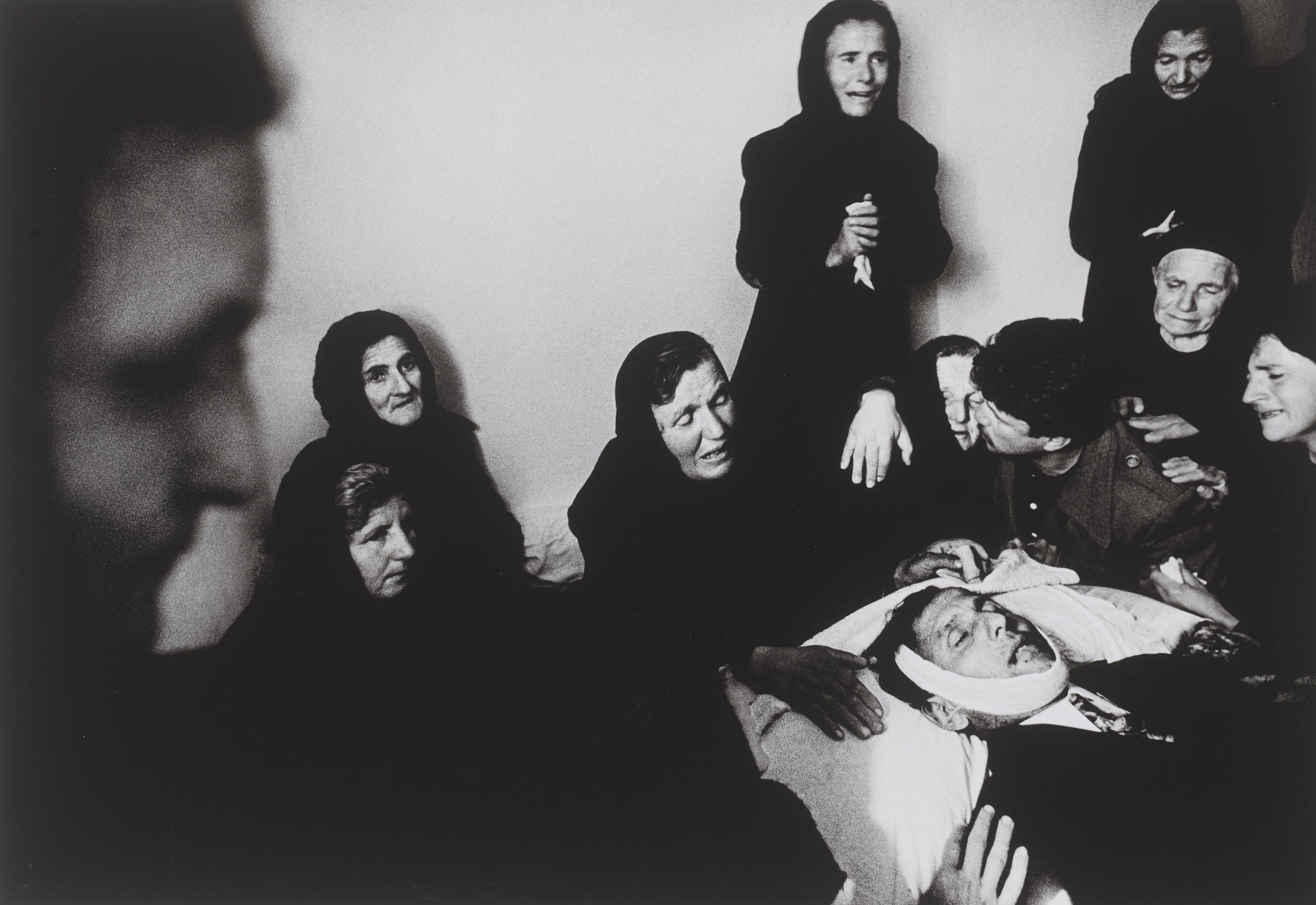 En gruppe kvinder iklædt sort græder over en mand på en båre med bandage om hovedet.