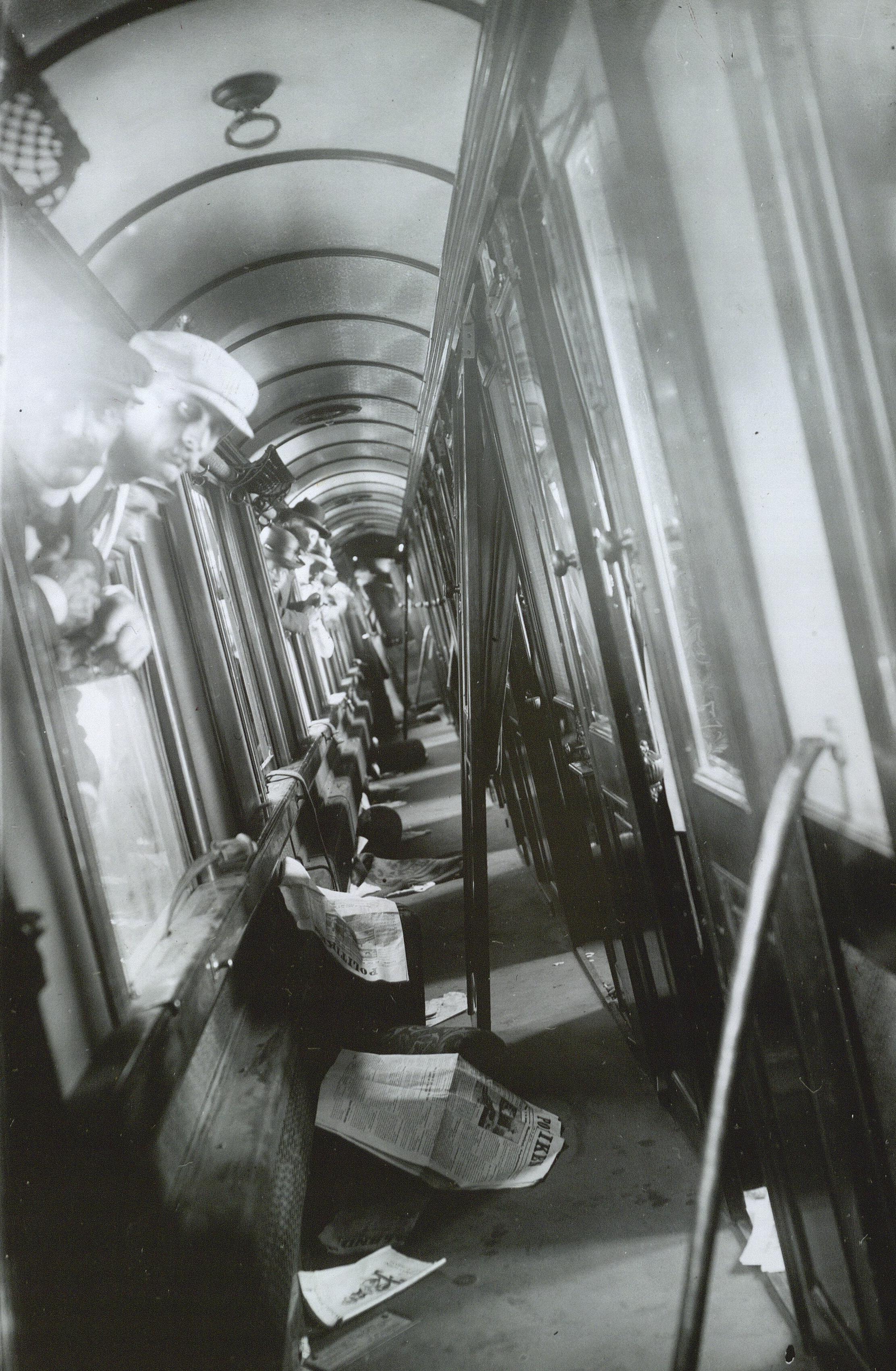 Flere mænd stikker hovedet ud af vinduer til gangen på et tog. På jorden ligger der aviser.