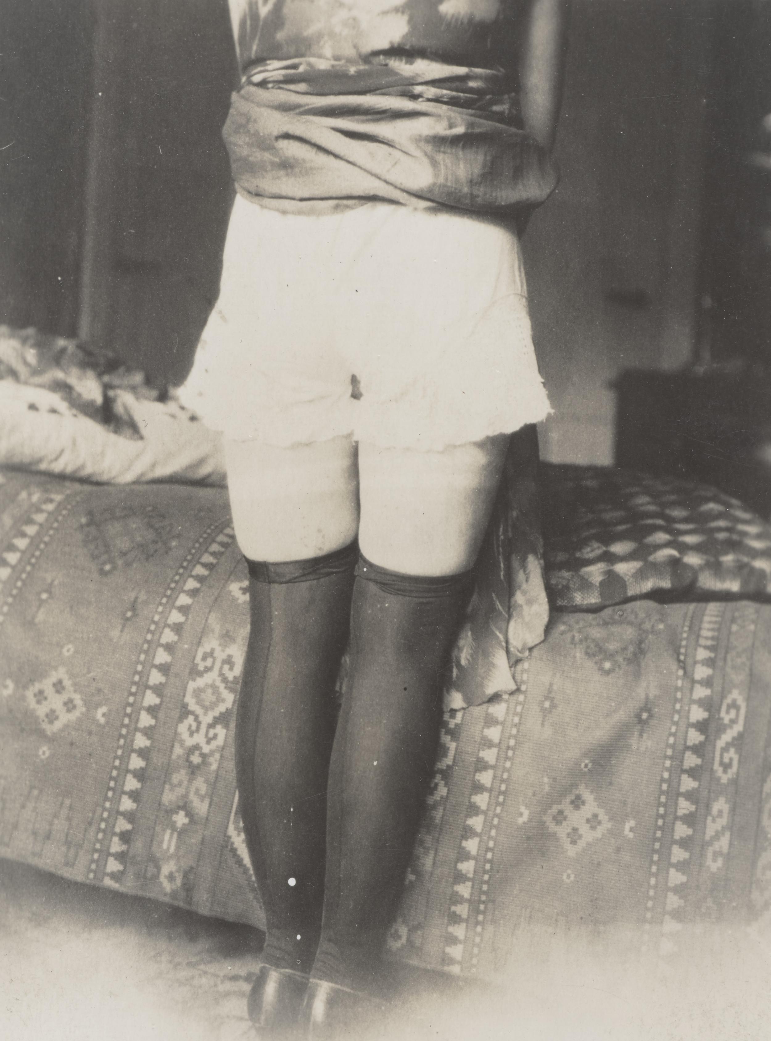Et par ben foran en seng. Der er hevet op i kjolen, så man kan se lange strømper og et par gammeldags underbukser