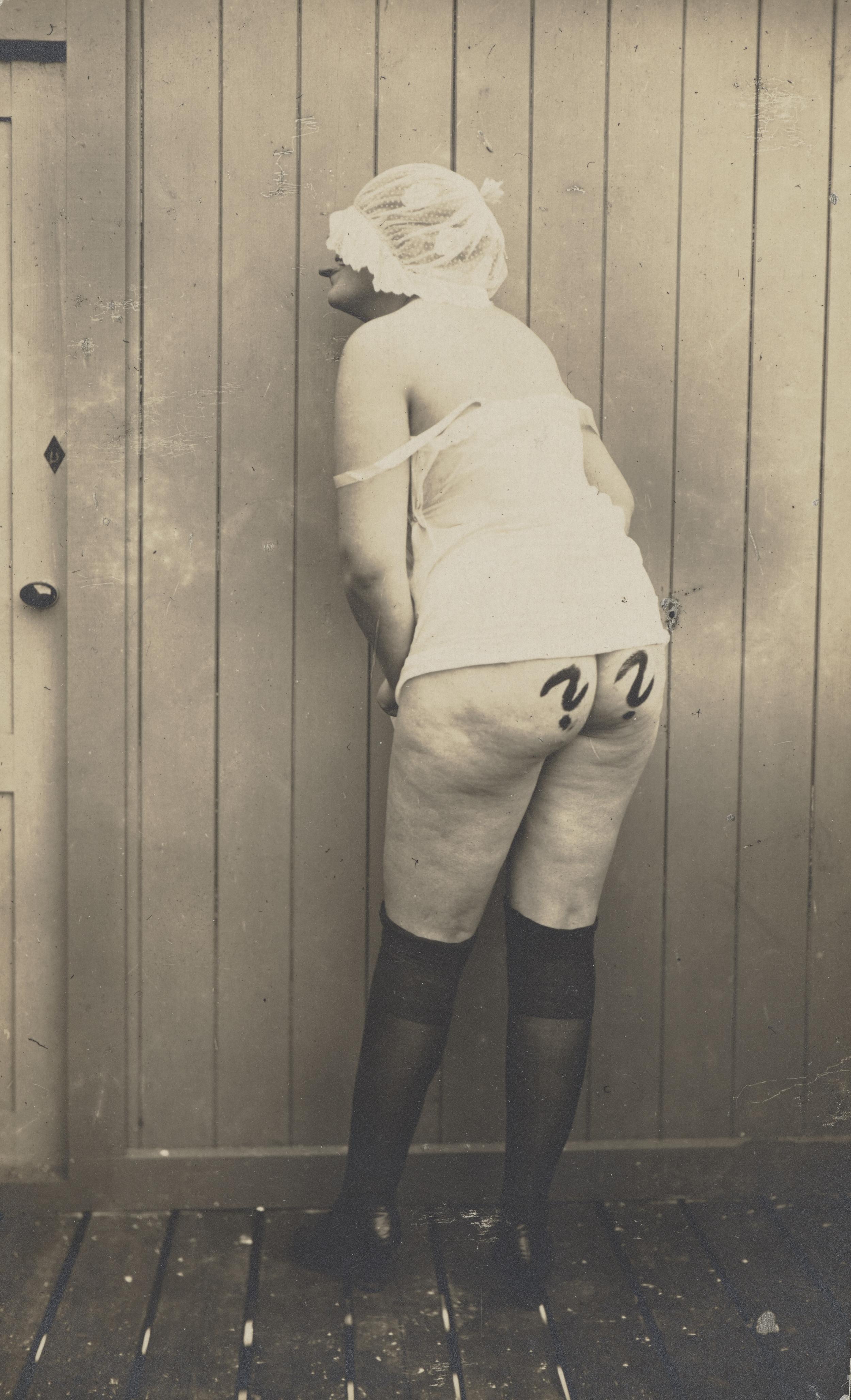 En kvinde med ryggen til kameraet iført kyse og lange strømper. Der er to spørgsmålstegn på hendes numse