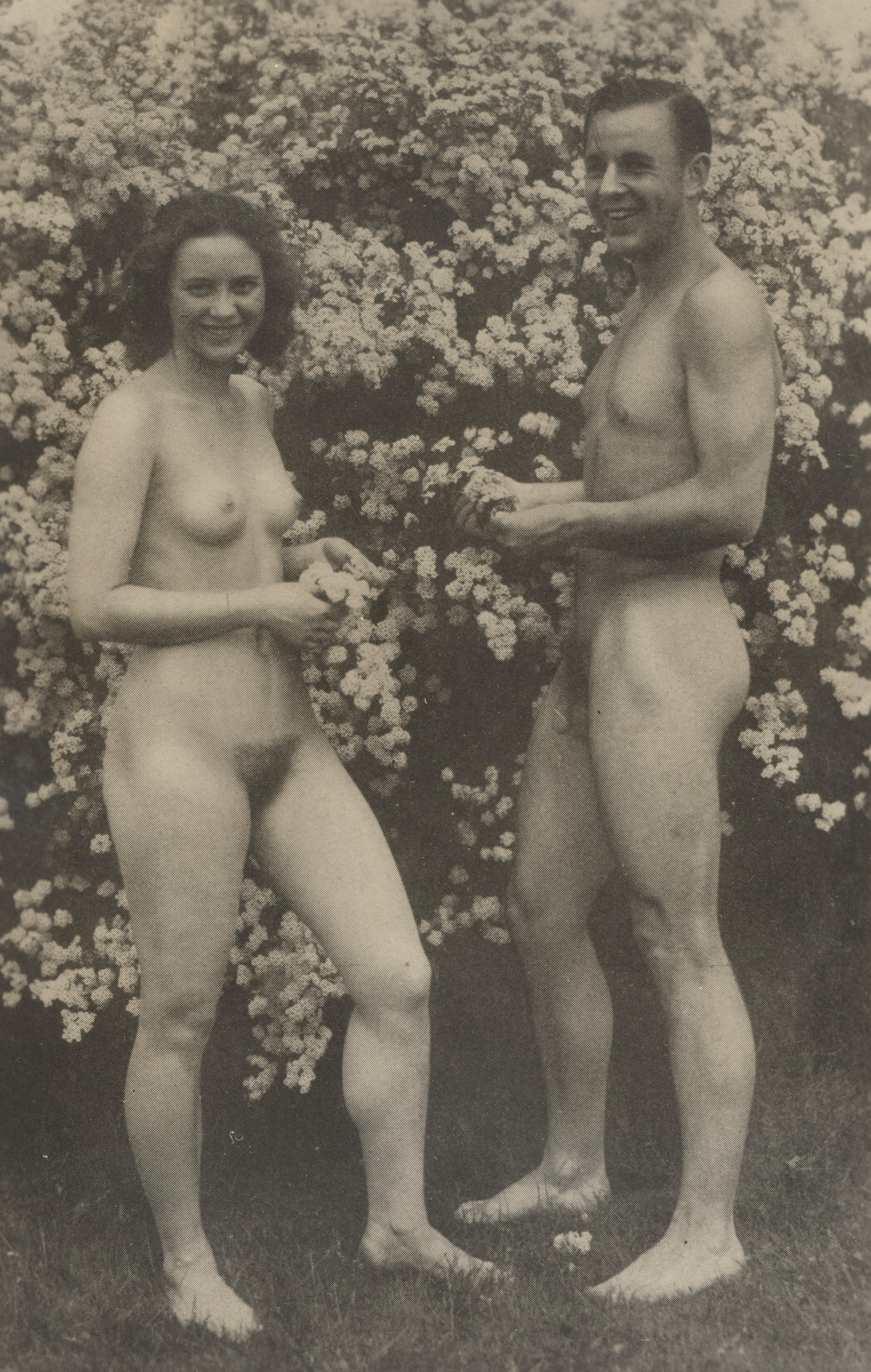 En nøgen kvinde og en nøgen mand står smilende foran en blomsterbusk