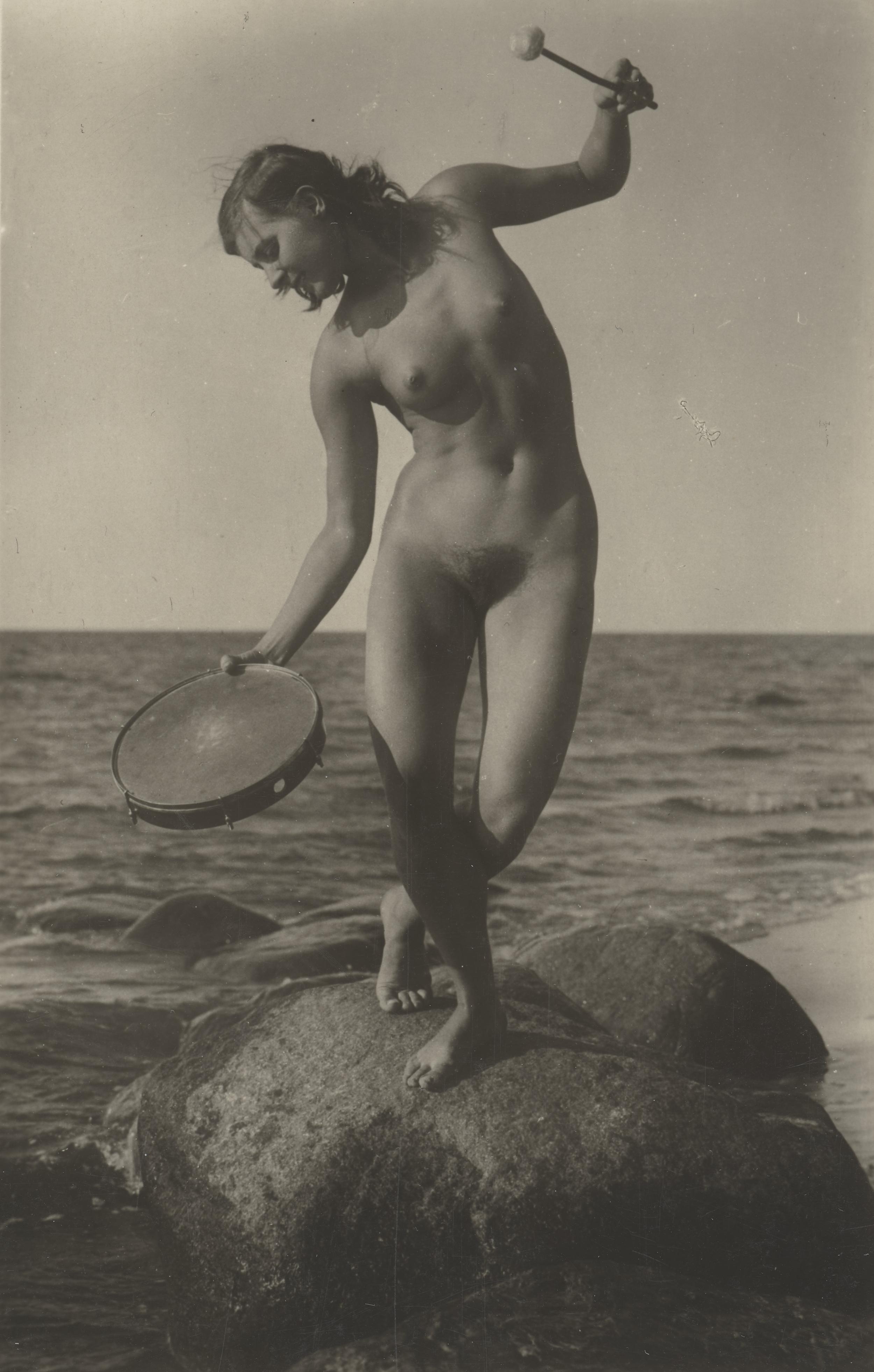 Nøgen kvinde på en sten ved havet med en tamburin i hånden