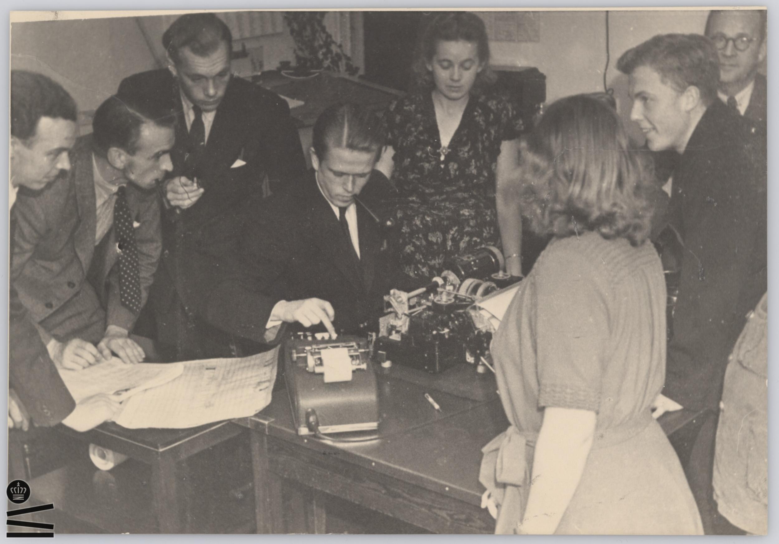 Man is bent over typewriter. Women and men watch.