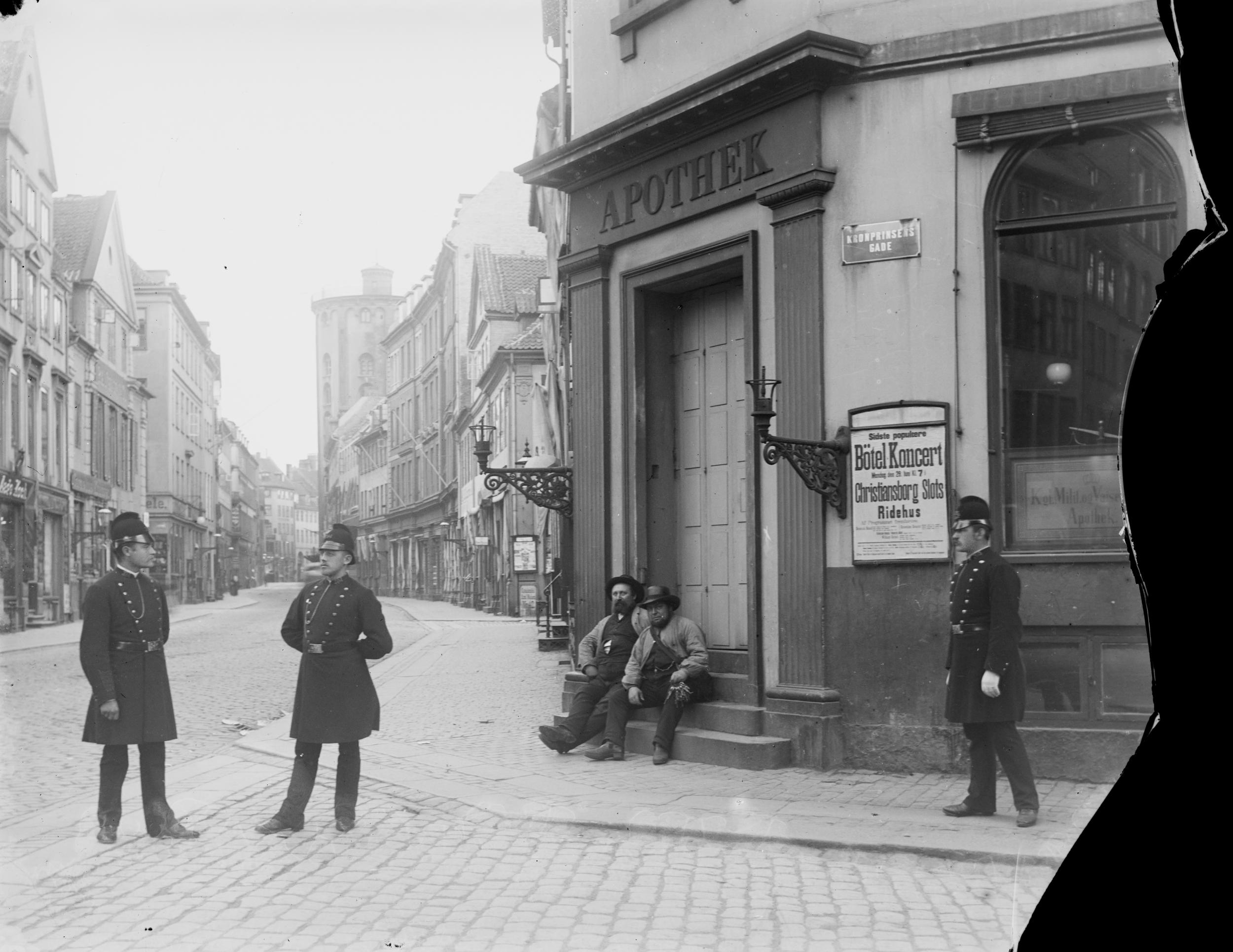 Tre politimænd står på et gadehjørne. To mænd sidder på trappen ved et apotek