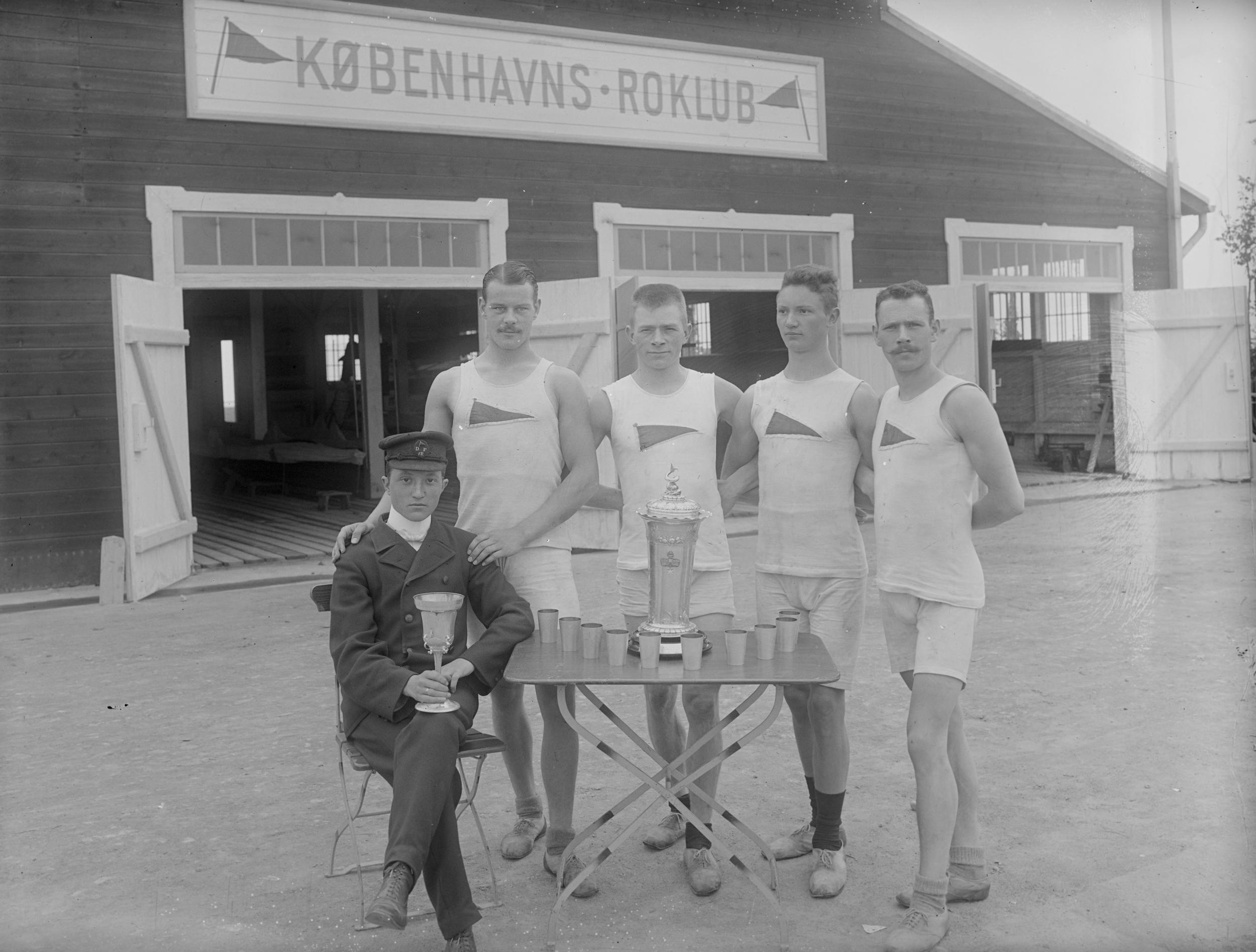 Fire mænd klædt i hvidt og en mand klædt i sort foran Københavns Roklub med pokaler