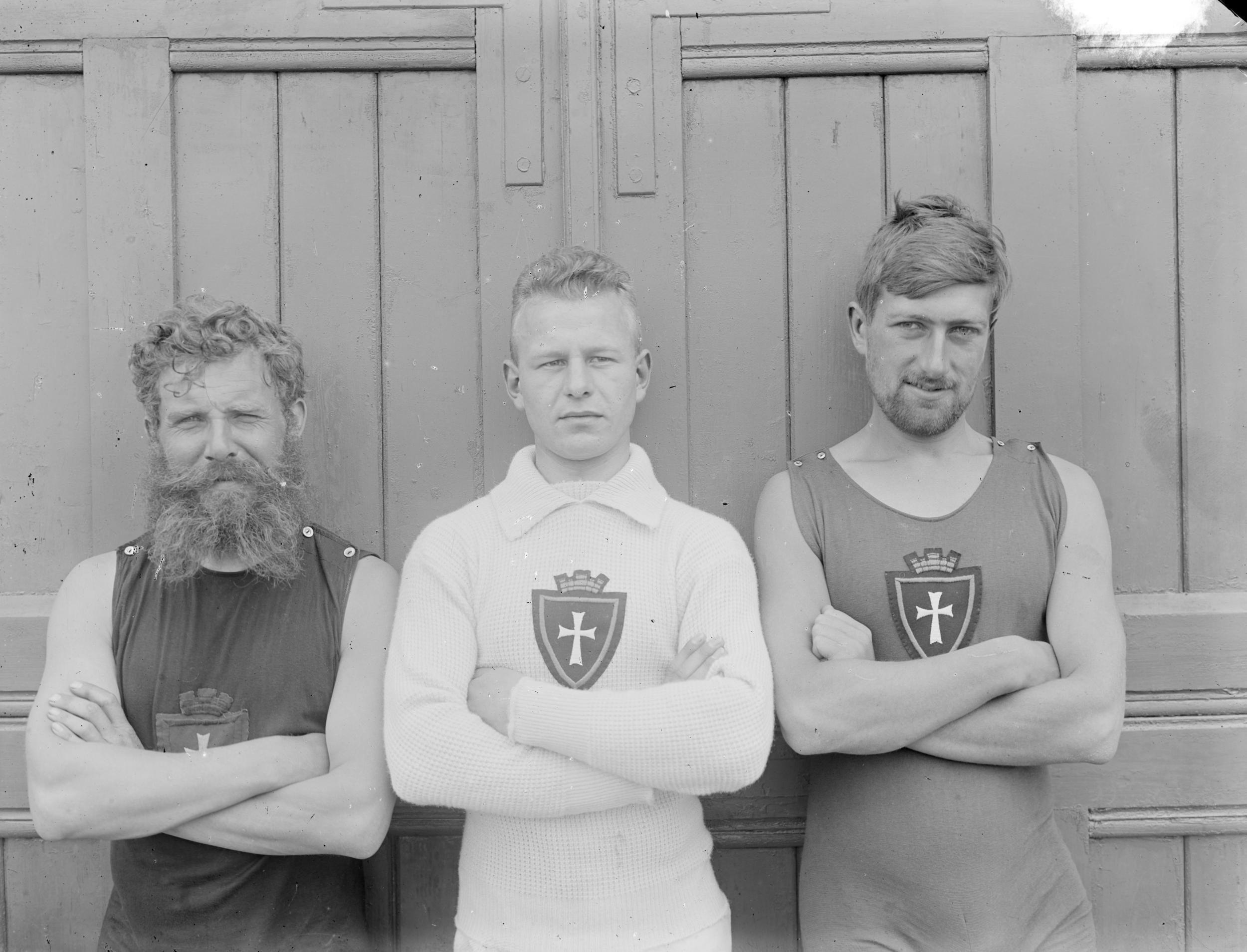 Tre mænd står med krydsede arme foran en dør