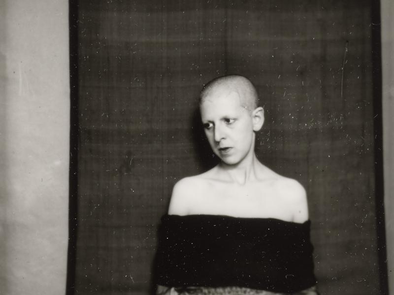 Selvportræt af Claude Cahun, der poserer foran et mørkt vægtæppe. 