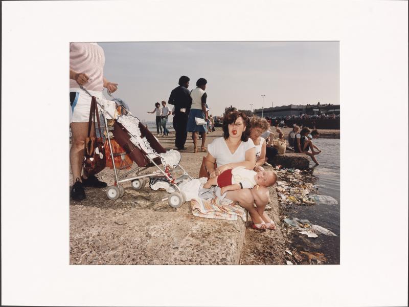 En kvinde sidder med en klapvogn og en baby ved noget vand, 1980erne